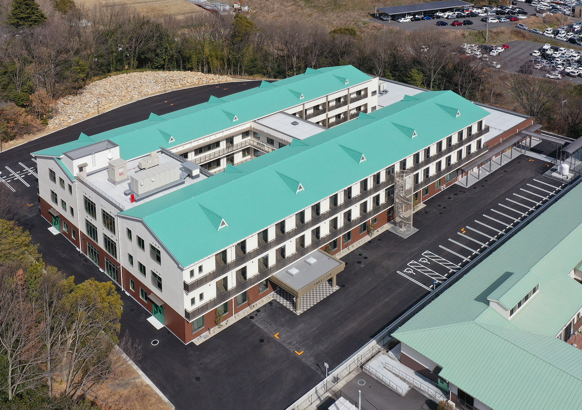緑屋根の介護施設建物の空撮。駐車場と周辺環境が確認できます。